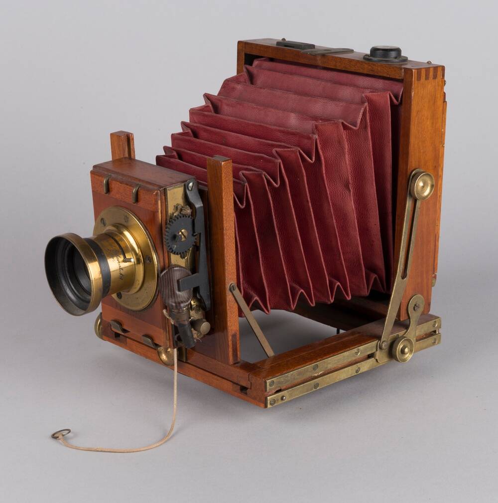 An old-fashioned camera stands against a plain grey background. It is mostly made from wood, with red leather bellows in the middle and a brass lens piece.