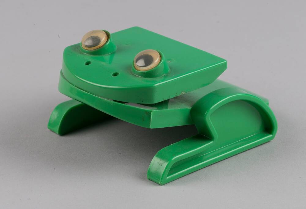 A bright green plastic paper holder, shaped like a frog, is displayed against a plain grey background. The paper would slot into its mouth. It has googly eyes on top of its head.