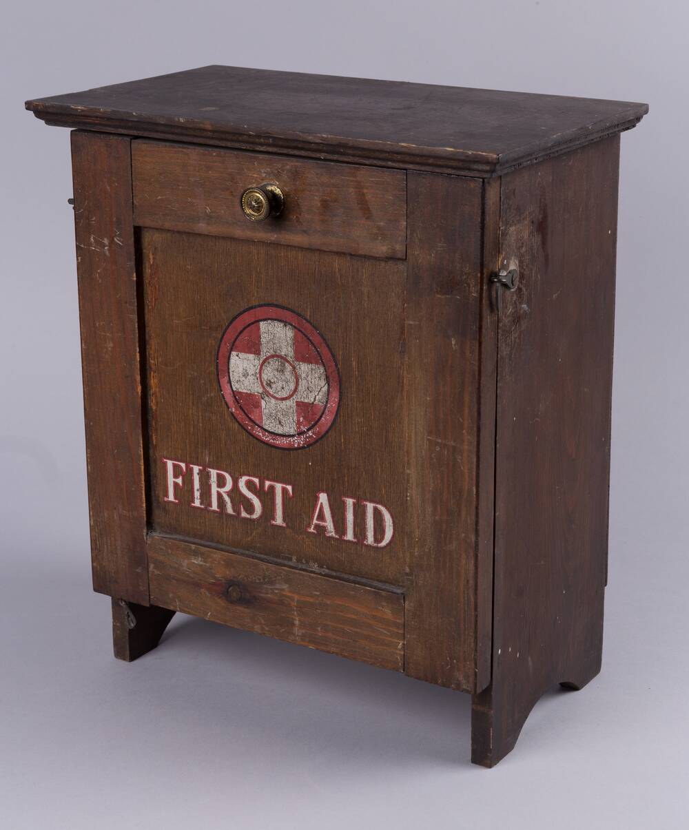 A small wooden cabinet has a white cross painted inside a red circle on the front door, above the words First Aid. There is also a small wooden drawer at the top with a brass handle.