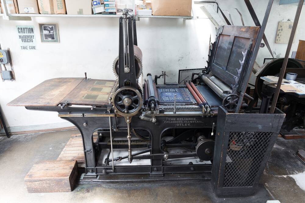 A very large printing press stands on a concrete floor in a printing works. Letterpress printed posters can be seen on the wall behind.