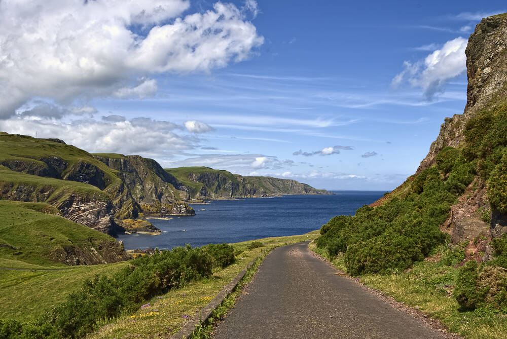 A narrow road leads down towards the deep blue sea, with a steep grassy cliff rising to the right. An indented rocky coastline can be seen ahead.