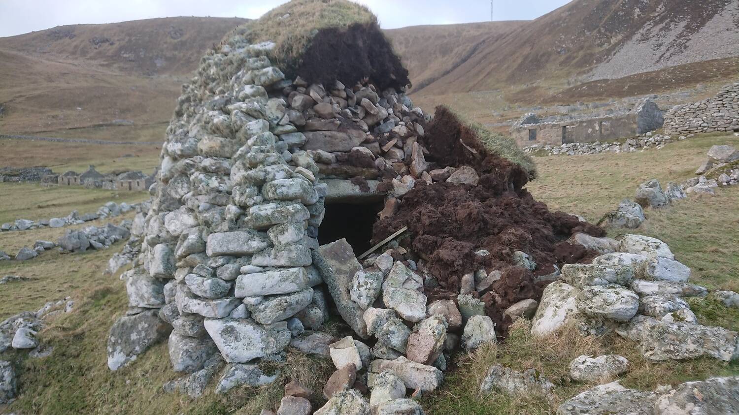A stone structure with turf roof. Some of the stones are lying around next to the structure. 