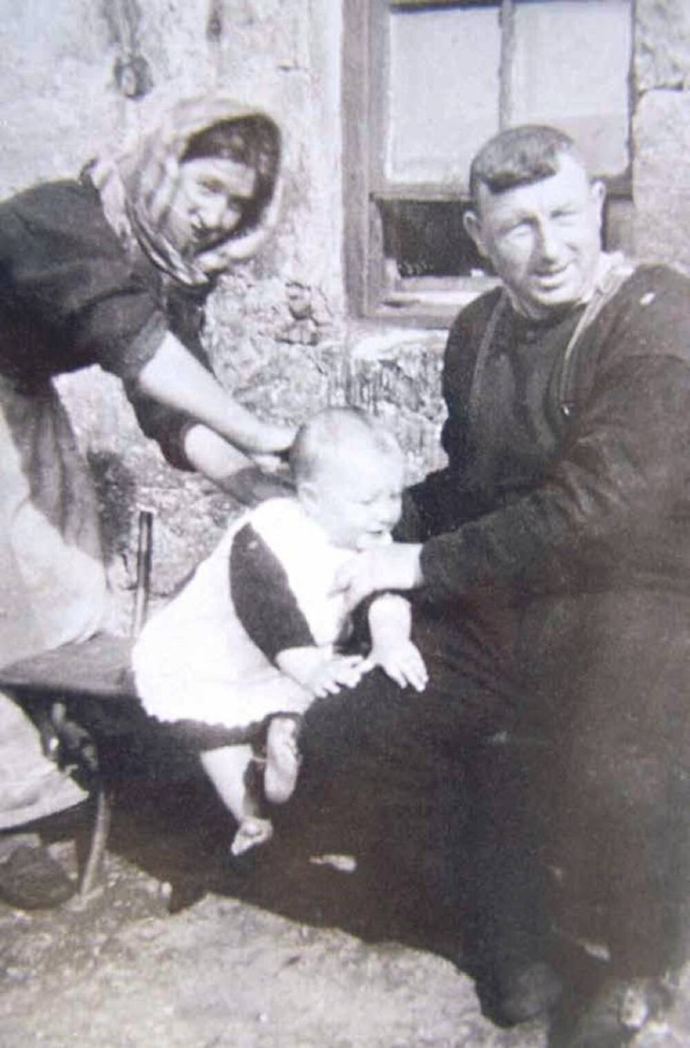 A black and white photograph of a young family sitting on a bench outside a stone cottage. The mother leans over a baby, who is perched on the bench and being held up by his father.
