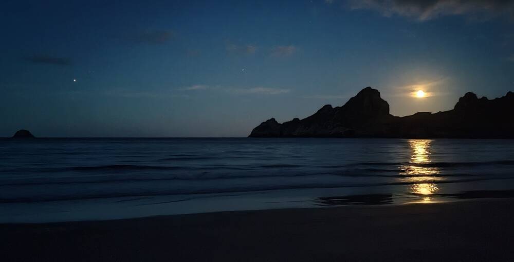 A night time view from the beach of Village Bay, looking out to sea and towards the jagged island of Dun. The rocks are silhouetted against the night sky. A full moon hangs in the gap between peaks, its light perfectly reflected in the still sea water.