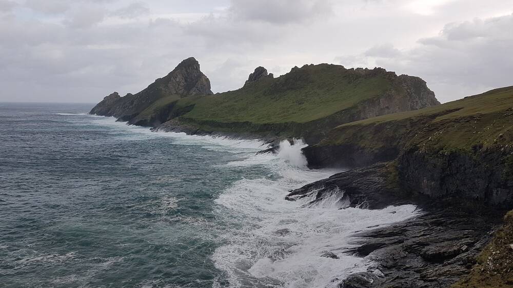 White foamy waves crash against the rocky sides of an island, where the jagged rocks fall straight into the sea. The water is a cold petrol blue; the sky is grey and heavy.