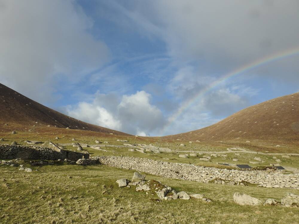 A rainbow arches over a gap between two hills on St Kilda. Grassy slopes and drystone walls can be seen in the foreground.
