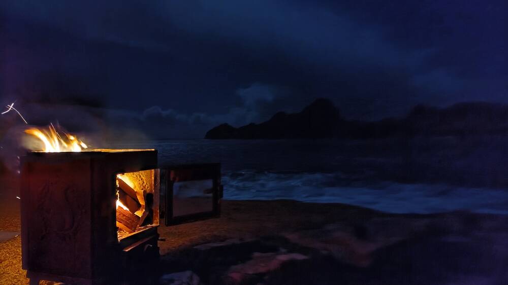 On St Kilda, an old wood burner converted into a brazier is lit and flaming. It's night-time, but just visible from moonlight are the sea waves and the island of Dùn in the background.
