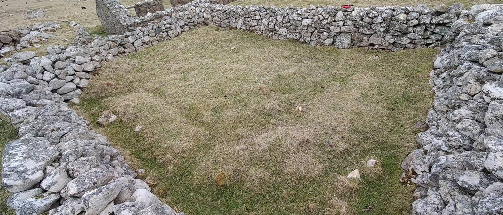 A dry stone wall surrounds the earthwork remains of a former structure, visible in the short stubby grass.