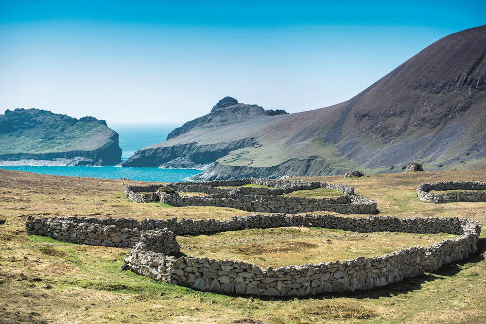 A view of Hirta in St Kilda, with the ruined remains of roundhouses in the foreground. Steep cliffs drop down to the blue sea in the distance.