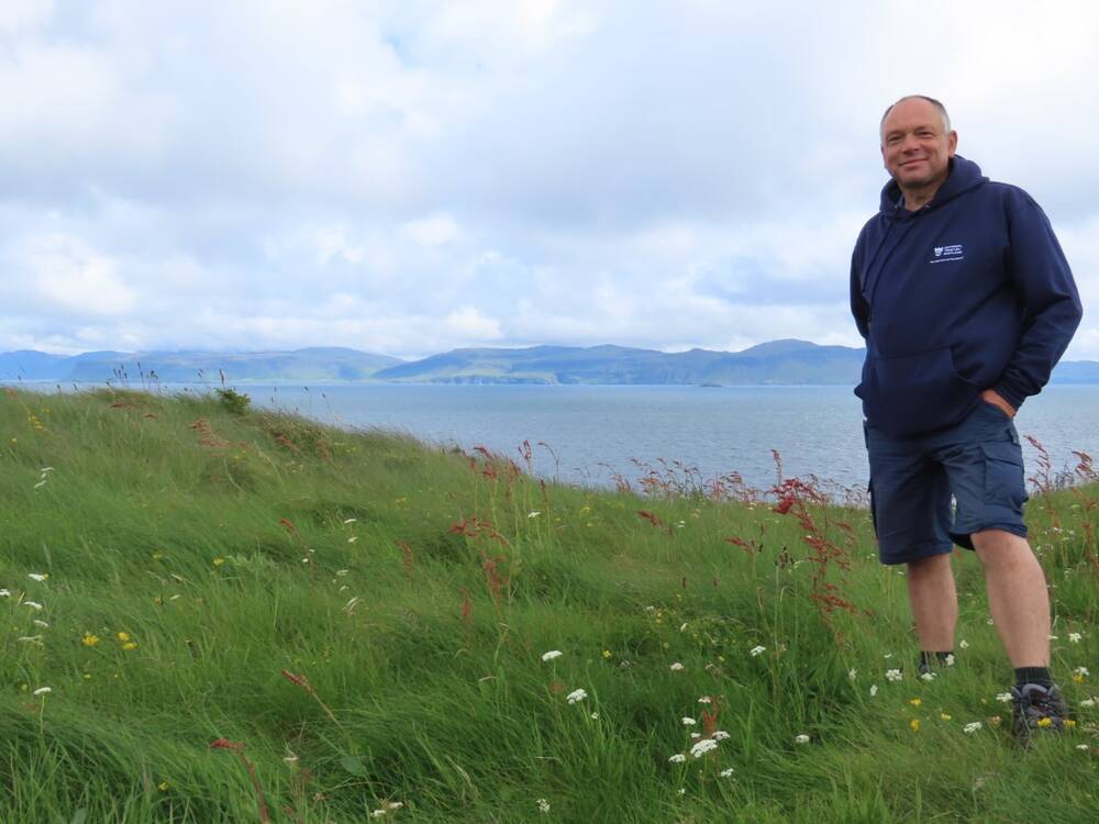 A man stands on a grassy cliff on Staffa looking towards the camera.