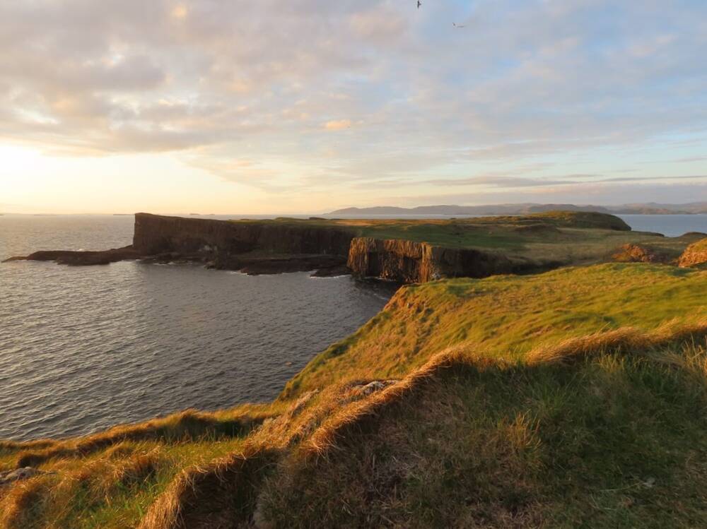 A view of the island of Staffa, as the sun sets over the horizon. The island has a very flat top, but almost vertical cliffs leading to the calm sea.