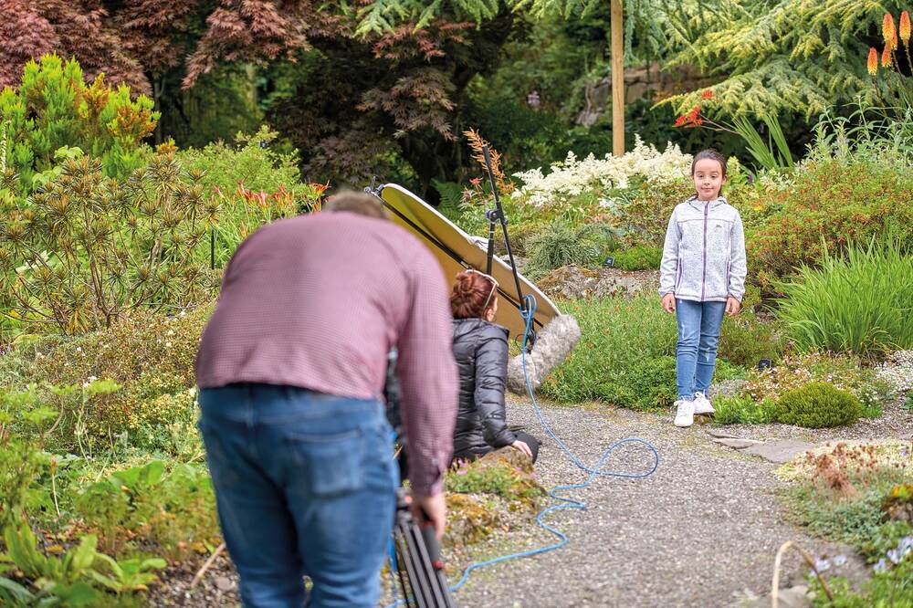 A young girl stands at the side of a path in front of a colourful flower bed, looking directly at the camera man who is filming in the foreground of this photo. A lady kneels on the path holding a light reflector.