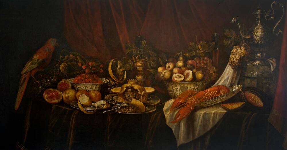 A long oblong-shaped oil painting of a still life of a richly laden dining table. Bowls are piled high with fruit; an elaborately constructed pie lies at the centre of the table; and a lobster is displayed to the right. A parrot sits on the left of the table. A jug shaped like a peacock stands on the right.