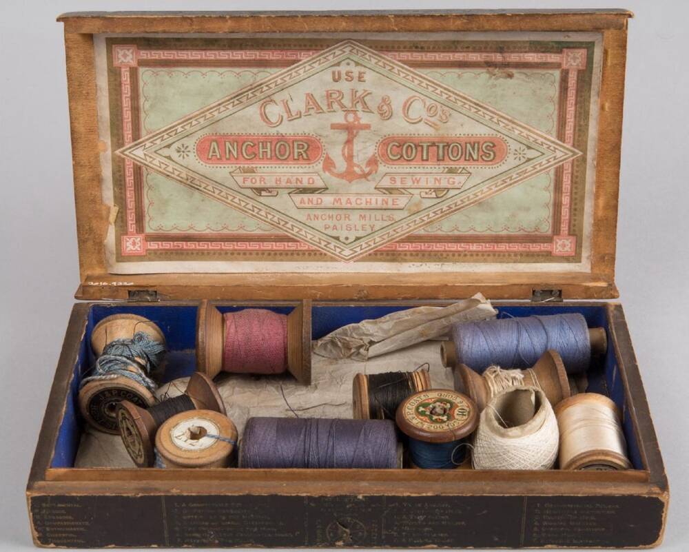 An old wooden box, open to reveal numerous cotton reels with thread of varying shades of blue, white, pink and black. The lower half of the box's interior is lined in blue material, while the top half displays a company label for Clark & Co.