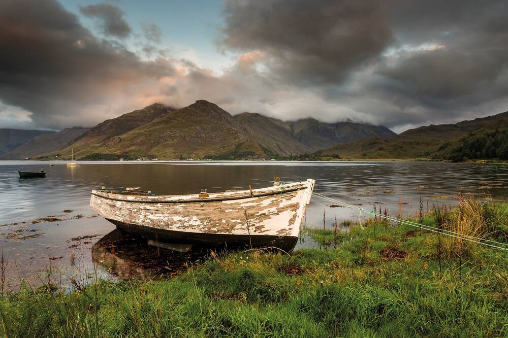A wooden rowing boat, with flaking white paint, is tied up at the edge of a loch beside some reeds. A couple of other small boats can be seen on the flat waters further out. Clouds, with glimpses of orange sunlight, hug the tops of the mountains in the background.