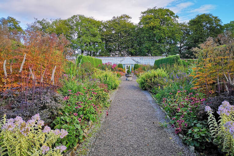 A wide gravel path leads through a walled garden towards a glasshouse at the far end. Either side of the path are beds filled with a wide range of plants and vegetables.