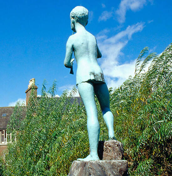 A green-tinged statue of Peter Pan stands on a rock pedestal, in the garden at J M Barrie’s Birthplace. It’s a sunny day, with a blue sky and bright green leaves on the nearby trees.