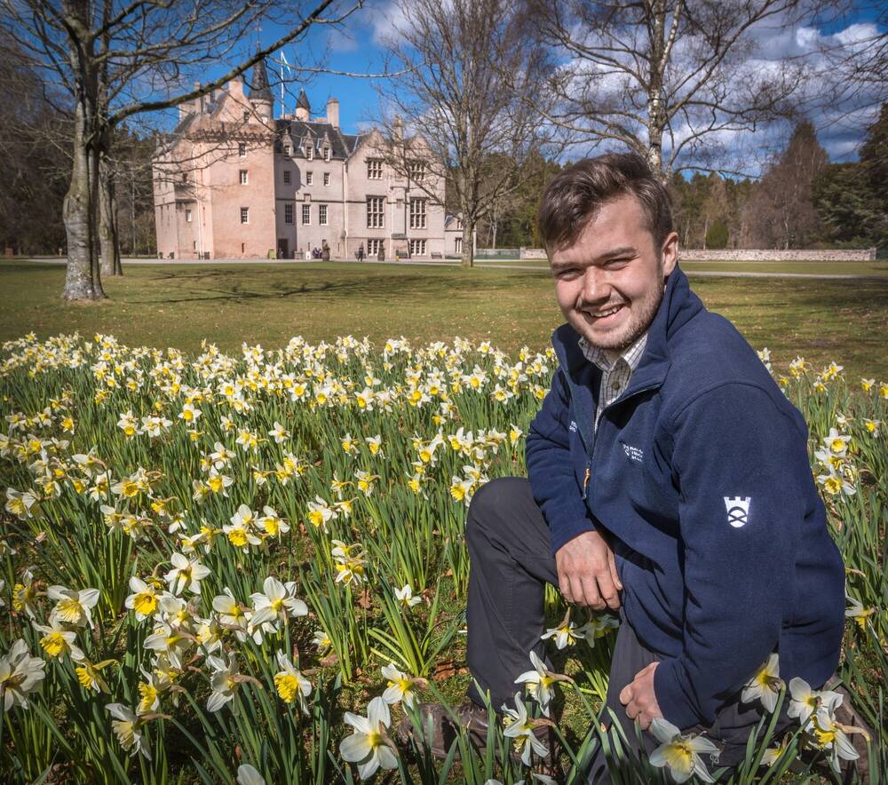  A smiling man in a  blue fleece is sitting among daffodils in front of Brodie Castle.