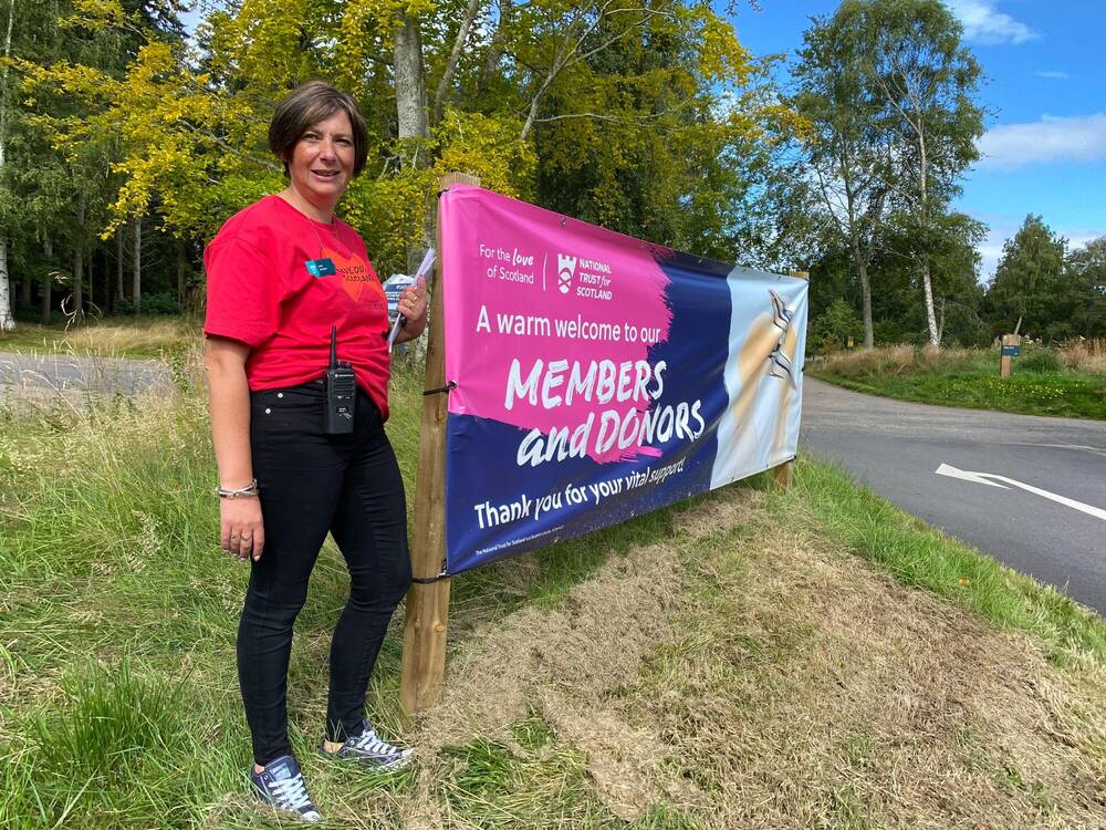 A woman is standing at a roadside next to a large National Trust for Scotland banner.
