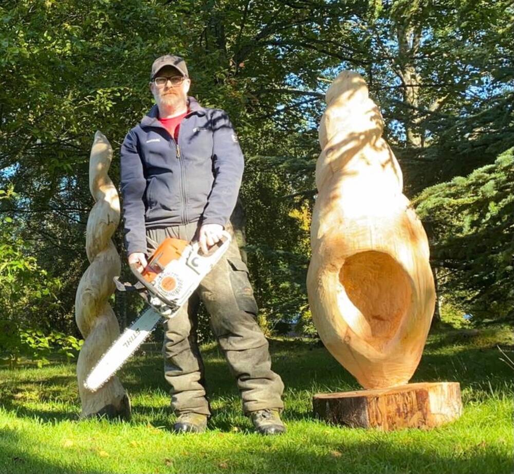 A man is holding a chainsaw next to two large wooden sculptures.