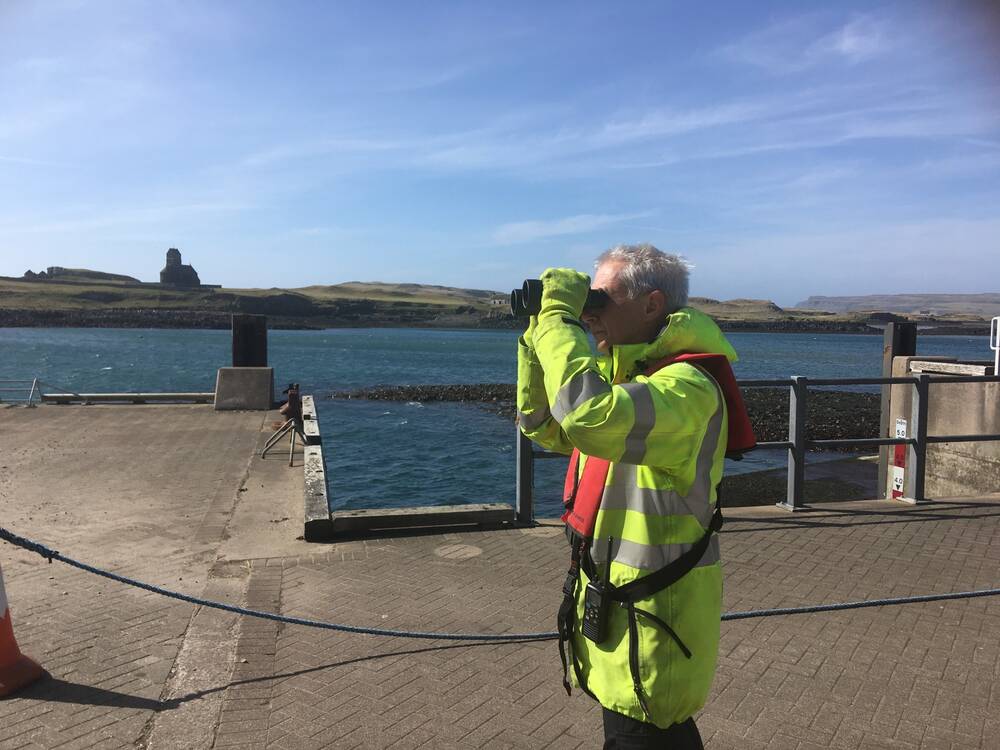 A man in a hi-vis jacket looks out to sea with binoculars while standing on a pier.