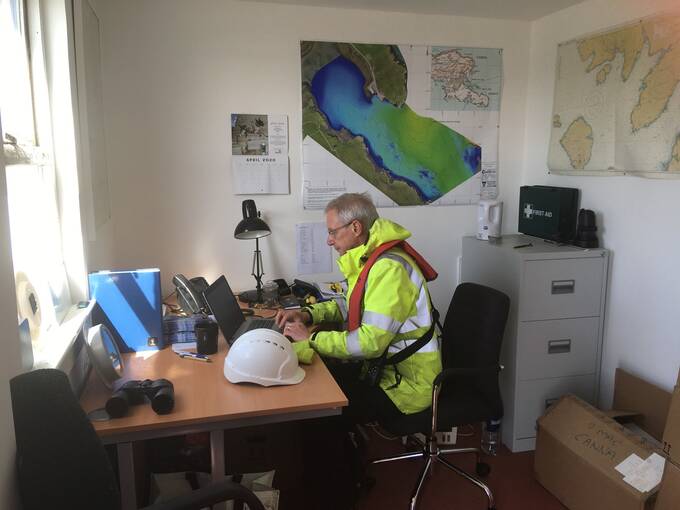 A man in a hi-vis jacket sitting at a desk looking at a laptop, in an office with shipping charts on the wall.