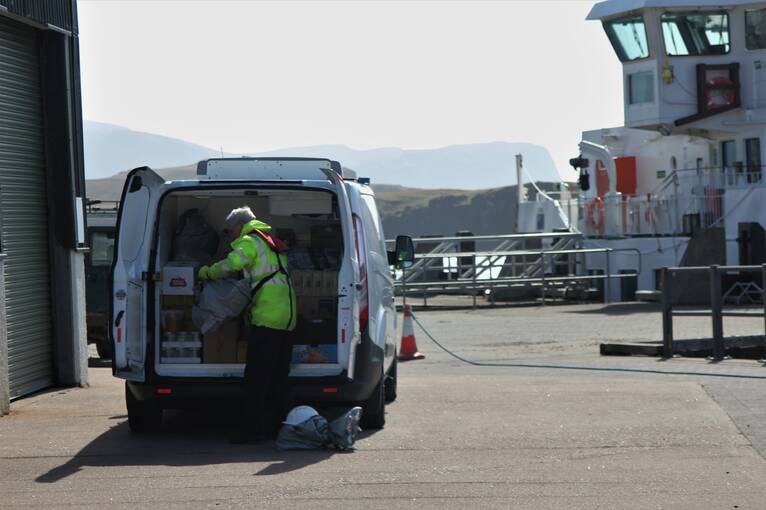 A man in a hi-vis jacket loading boxes into the back of a van, with a ferry in the background.