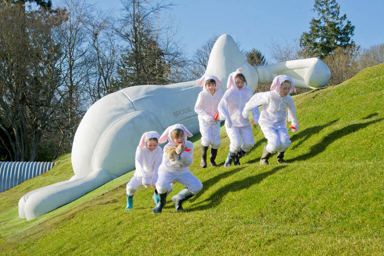 Brodie Bunny, a giant 6.5m high white rabbit, relaxing on the grassy slopes with children dressed in rabbit suits playing