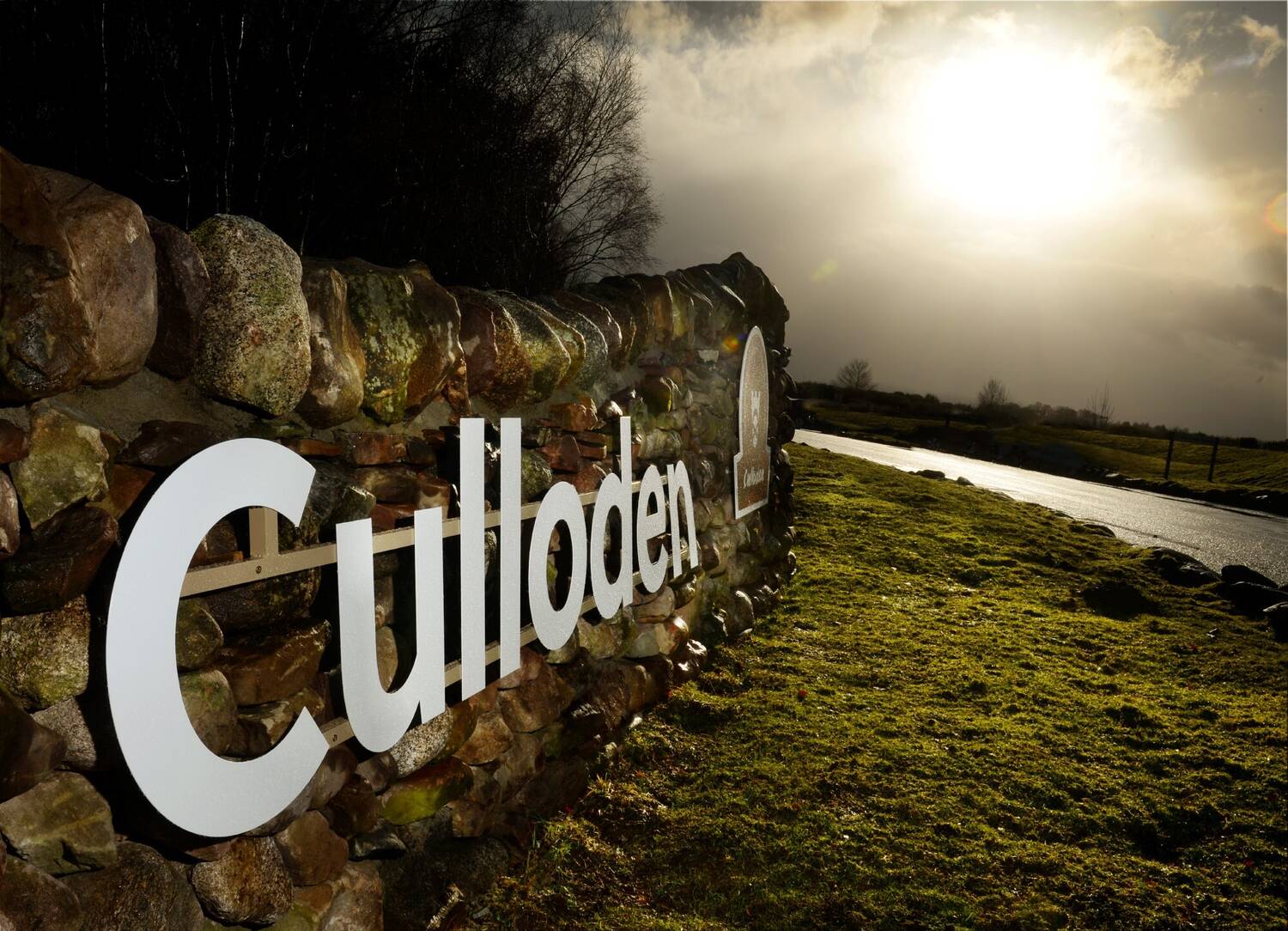 Metal letters spelling out Culloden are attached to an old stone wall at the entrance to a driveway. It has rained recently and the grass and stones glisten as the sun breaks through the clouds.