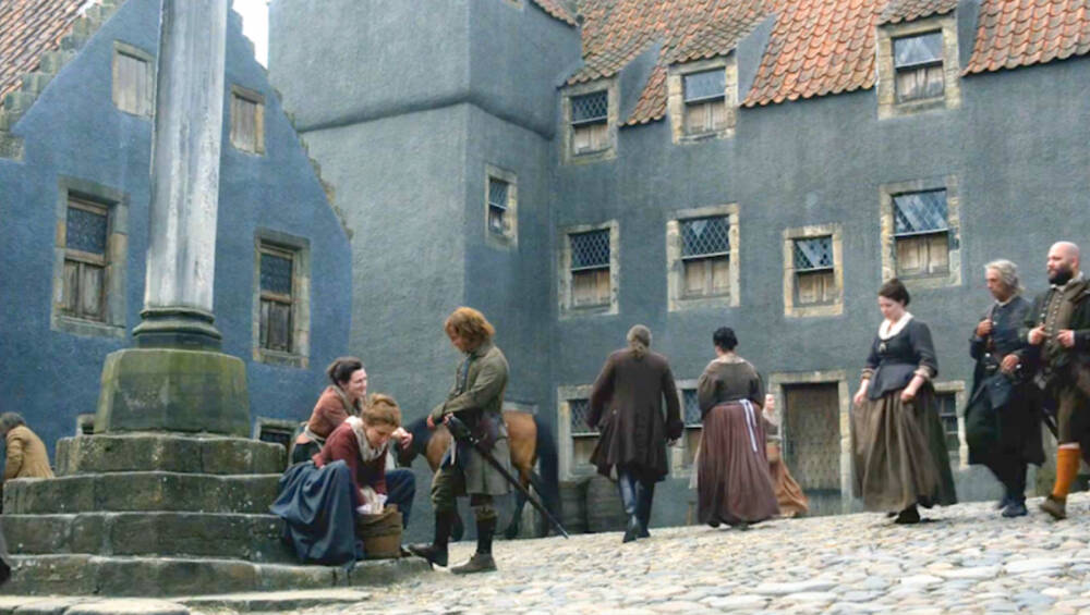 A scene from Season 1 of TV series Outlander showing people in 18th-century clothing milling around the cobbled village square. The buildings are coloured a washed-out blue.