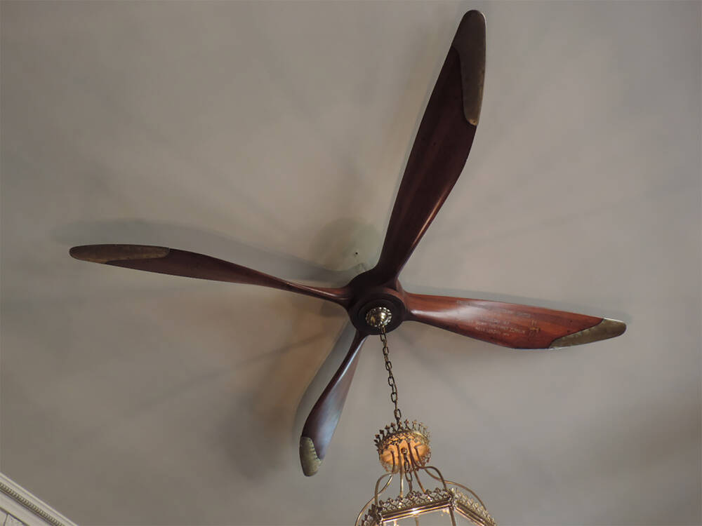A large wooden aeroplane propeller, with brass tips, is attached to a ceiling. From it hangs a brass chain, holding an elaborate light fitting.