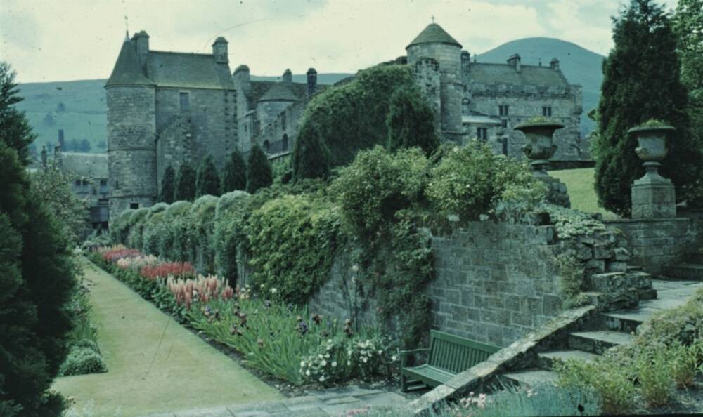 A photograph taken in the 1950s shows a palace in the background, with cypress trees in front of a ruined range, and a colourful flower bed below.