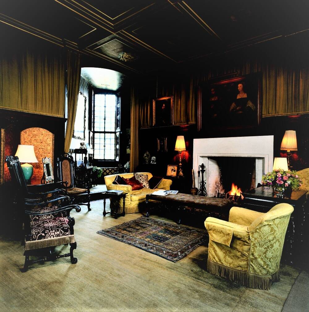 A drawing room with dark panelled ceiling and hessian-lined walls. There is a large fireplace with a lit fire.