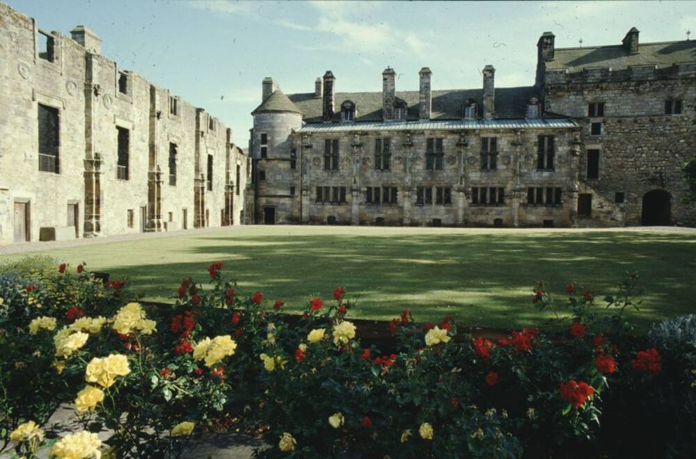 A rose  bed with yellow and red roses, with a ruined part of a palace to the left and a roofed range behind it. The photo was taken in the 1950s.