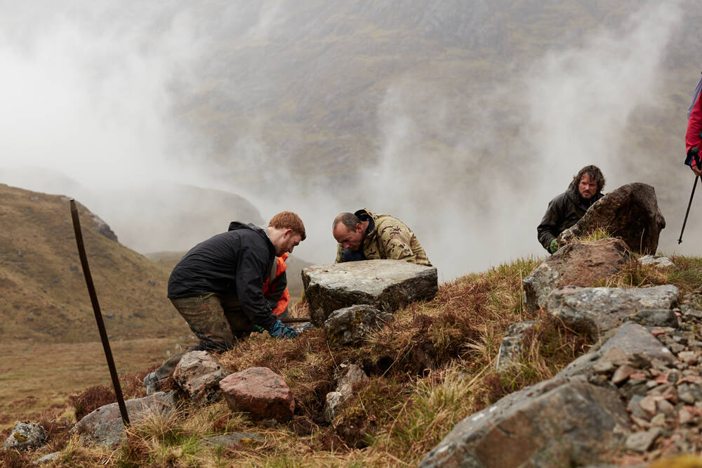A group of men at work on a high mountain footpath, as mist swirls in the background. Three are levering a large boulder into place.