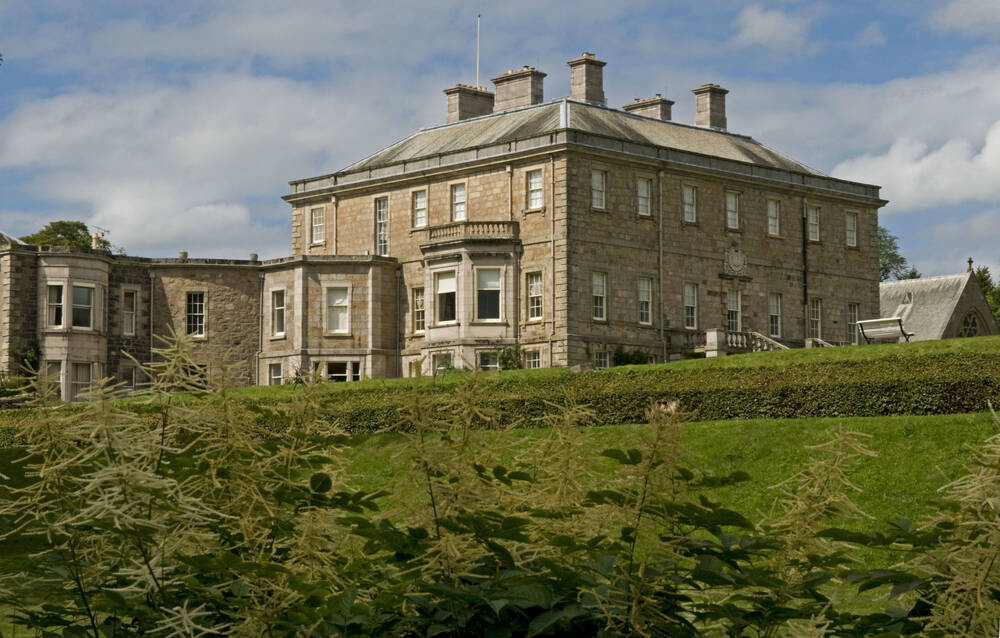 A view of a grand mansion house, with a classical Georgian exterior, seen from below in a terraced garden. Tall leafy plants grow in the foreground.