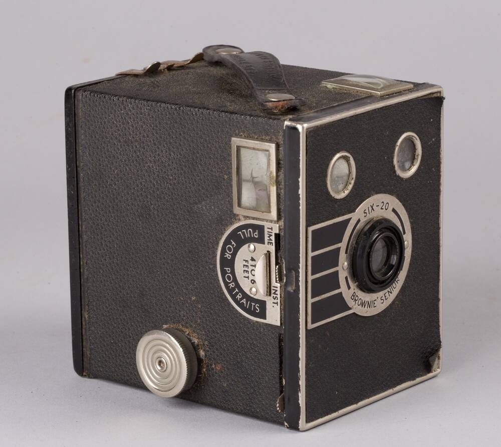A small, black, box-shaped camera with a leather handle on the top, a metal dial on one side and a lens at the front.