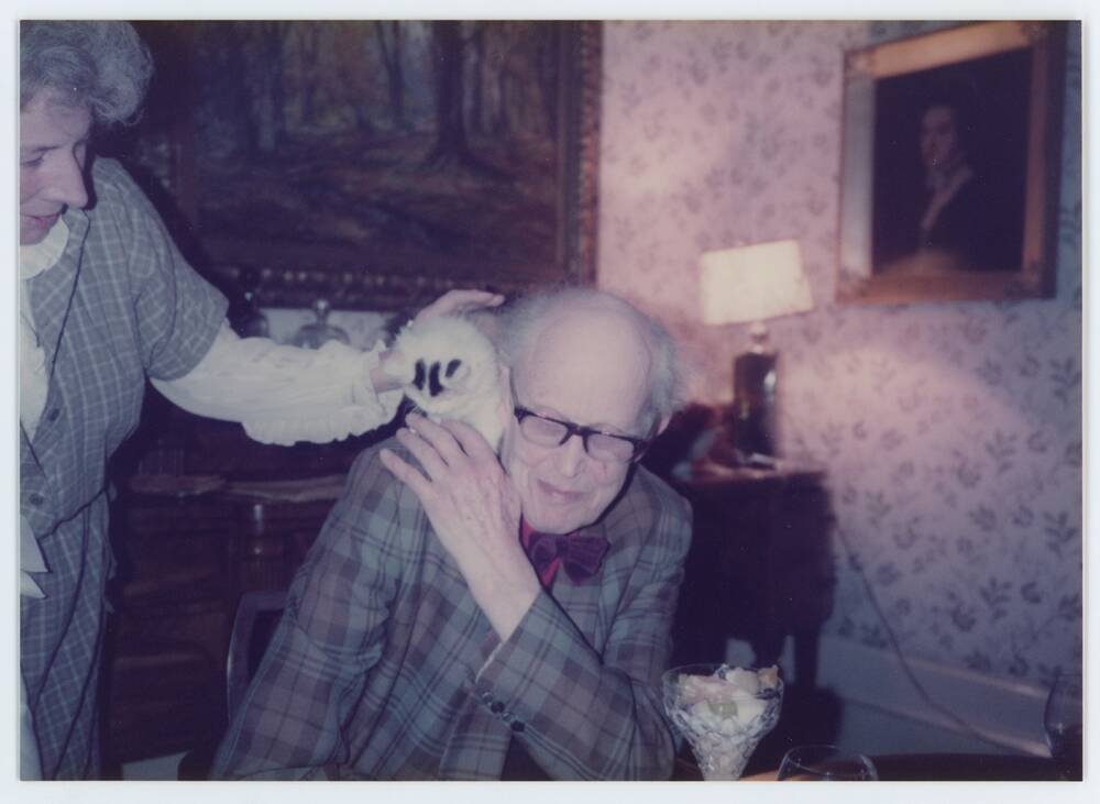  A colour photo of an old man inside, with a tortoiseshell cat on his right shoulder. A woman to his right is stroking the cat.