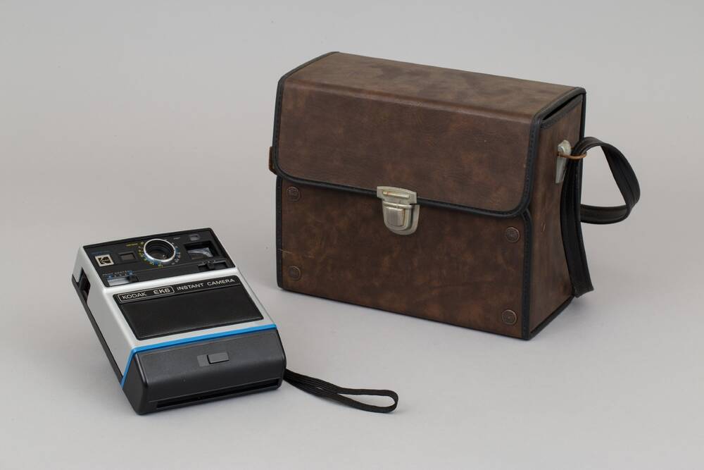 A rectangular, black and white plastic camera. The lens and print controls are visible. It lies next to a brown leather case.