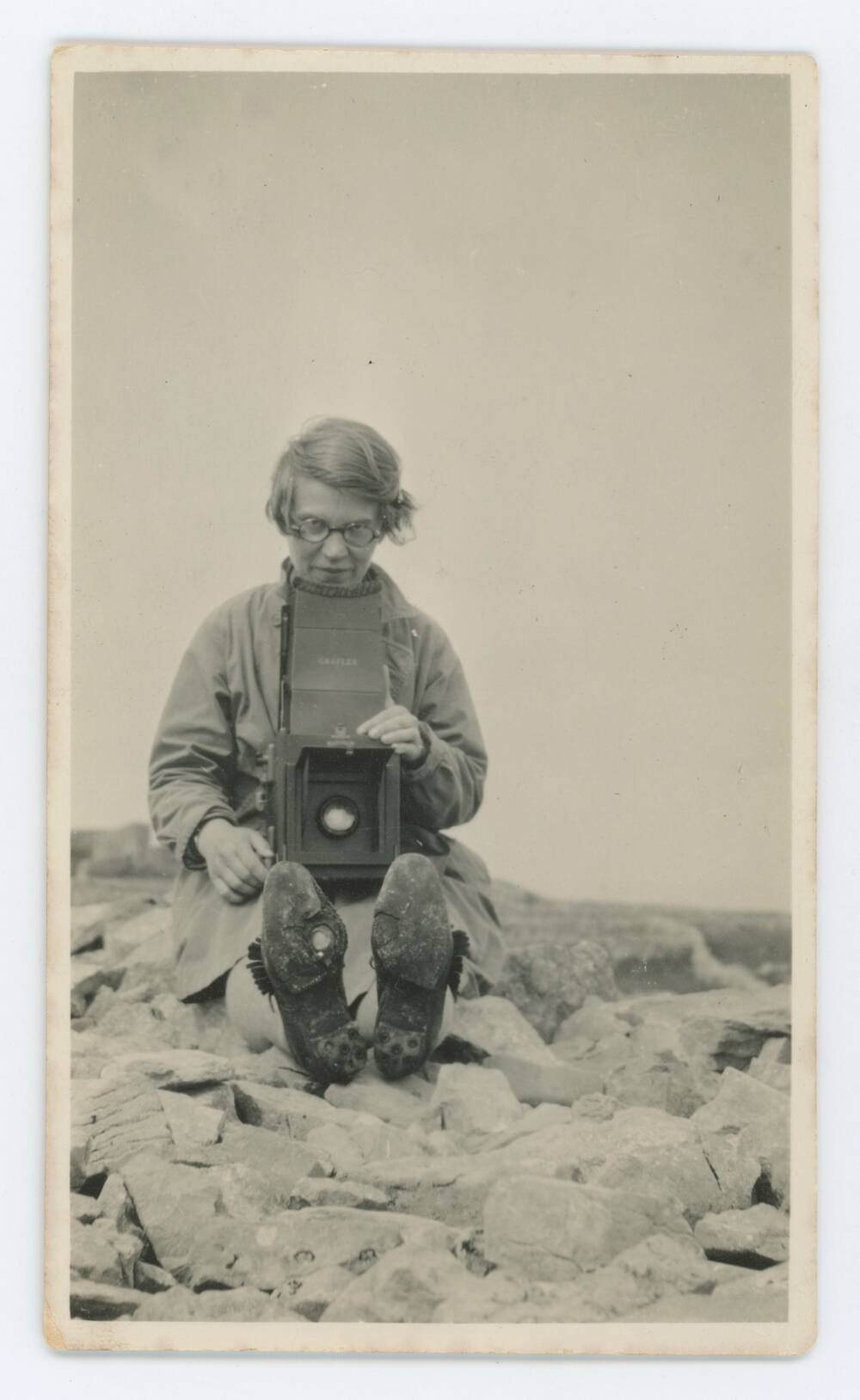 A black and white photo of a woman sitting on a rocky beach with her boot soles facing us. She holds a camera on her lap.