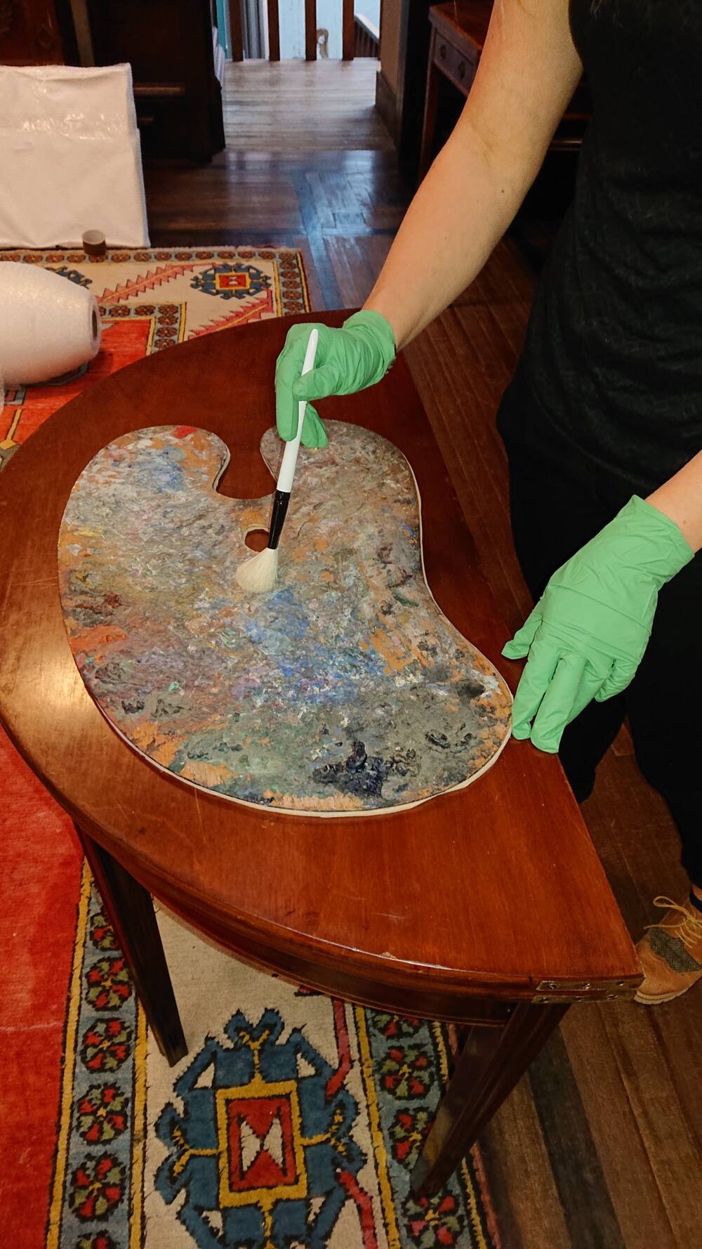 A painter’s palette on a wooden table. Someone wearing green latex gloves is carefully using a brush to clean the surface of the palette.