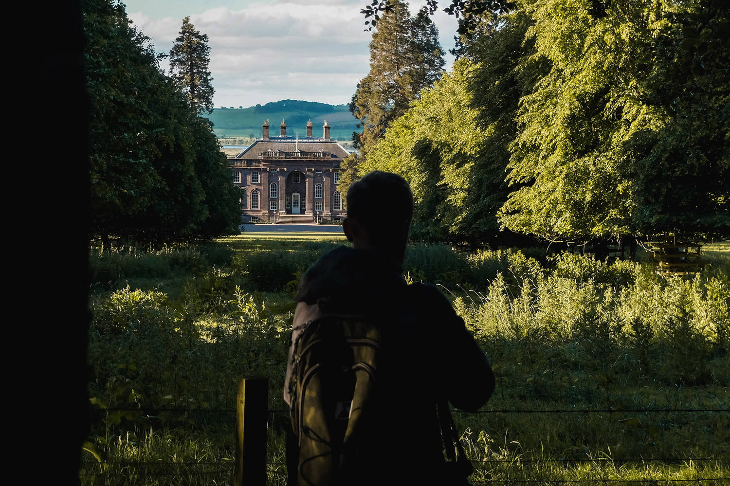 A man with a backpack on looks across a sloping grassy field, flanked by a variety of trees in full leaf. At the bottom of the field is House of Dun. The sun is rising over the House and field, and the man is in shadow while the landscape is cast in early golden sunlight.