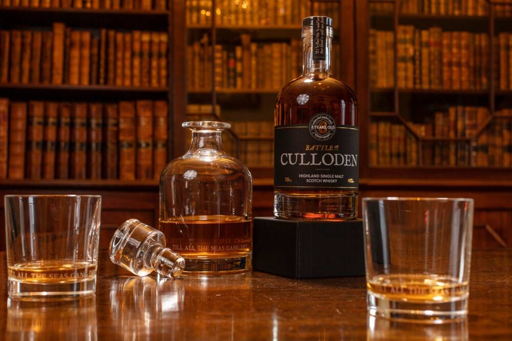A bottle of Culloden whisky sits on a table, with a decanter of whisky and two glasses of whisky. A bookcase with old books is the backdrop.