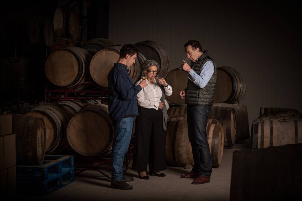A woman and two men are sampling whisky, standing in front of whisky barrels on their side.