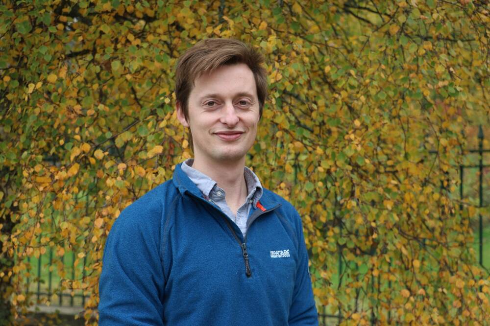 Head and shoulders of a man in a  blue fleece. He is standing in front of autumnal trees.