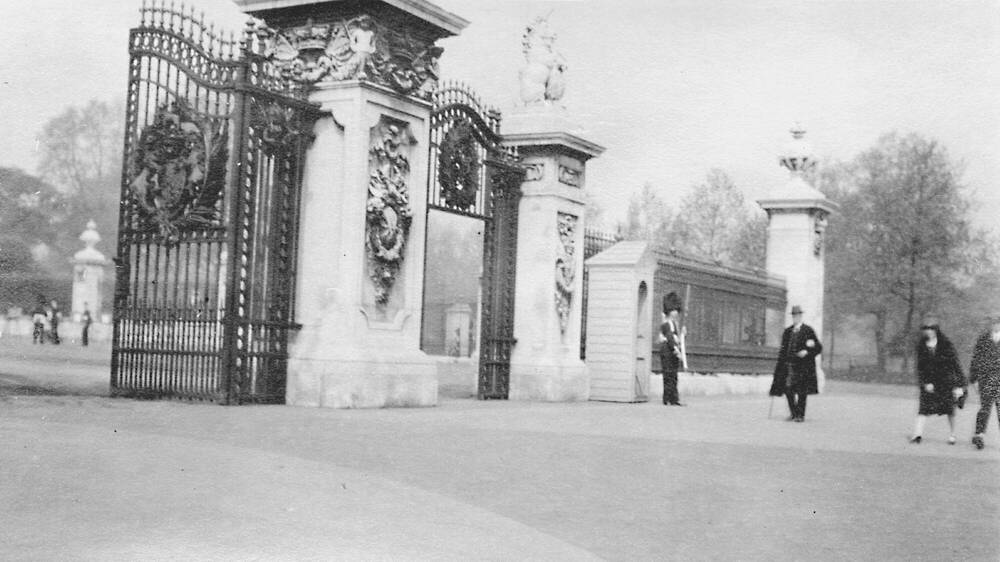 Black and white photograph from the 1920s of the gates of Buckingham Palace, with a few people walking past.