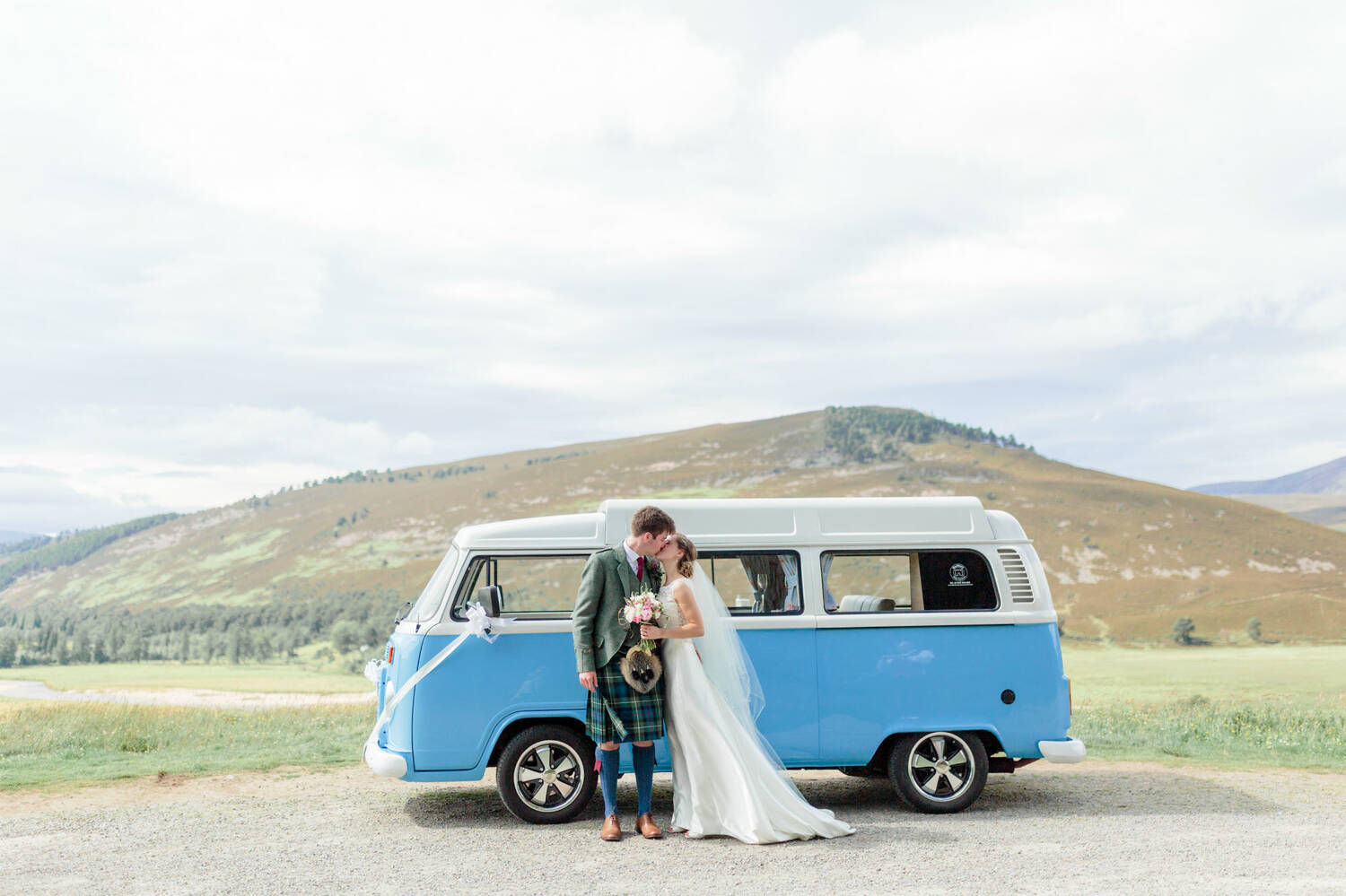 A bride and groom kiss in front of a blue camper van, parked in front of a mountain and moorland.