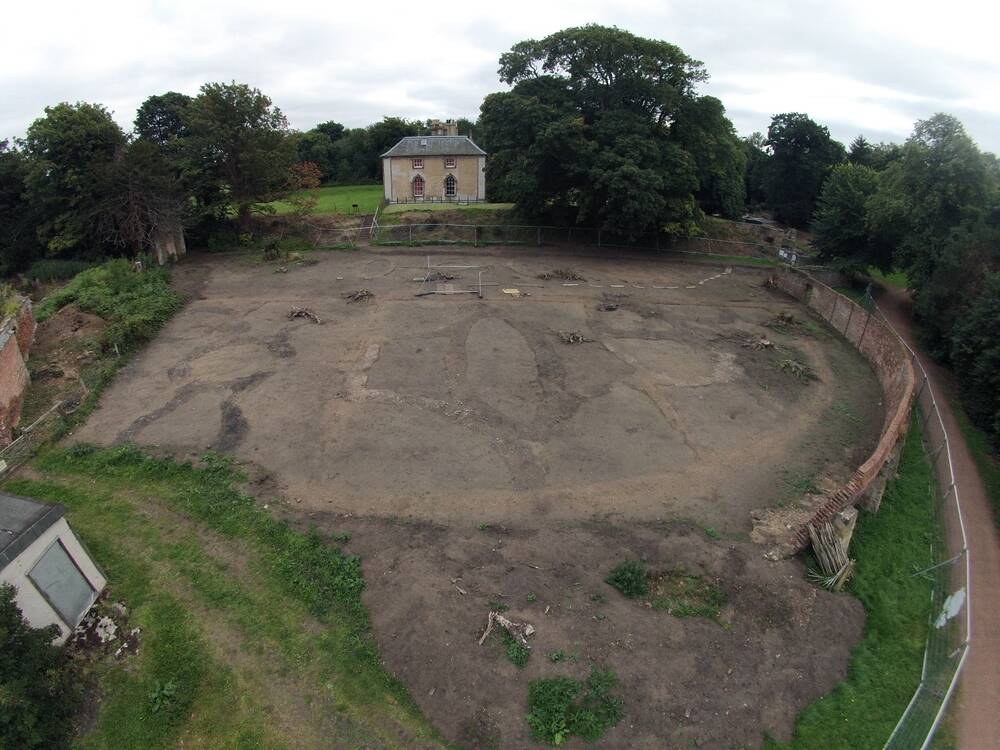 Bird's-eye view of a cleared area of ground being archaeologically investigated, with a house in the background, surrounded by woodland.