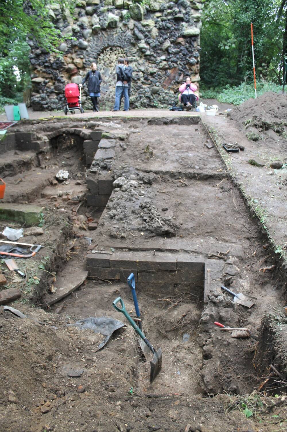 Archaeological excavation in woodland in front of a grotto. In the trench are the remains of walls and ditches,