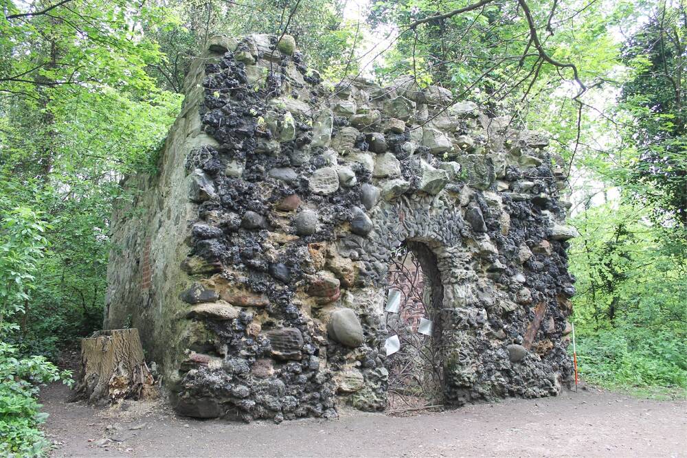 A shell house grotto in woodland. The walls are constructed of large boulders and there is a central doorway.
